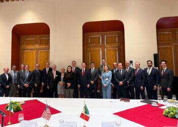 Group of professionals in business attire posing for a formal group photo in a conference room with large wooden doors in the background, seated and standing behind a long table with place settings.