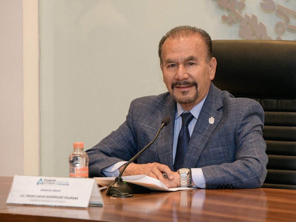 Smiling man in a blue checkered suit speaks at a conference table with a microphone in front and a nameplate reading Lic. Pedro David Rodríguez Villegas on the desk.